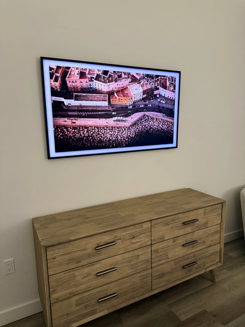 A Frame TV showing aerial art, mounted on a beige wall above a wood dresser