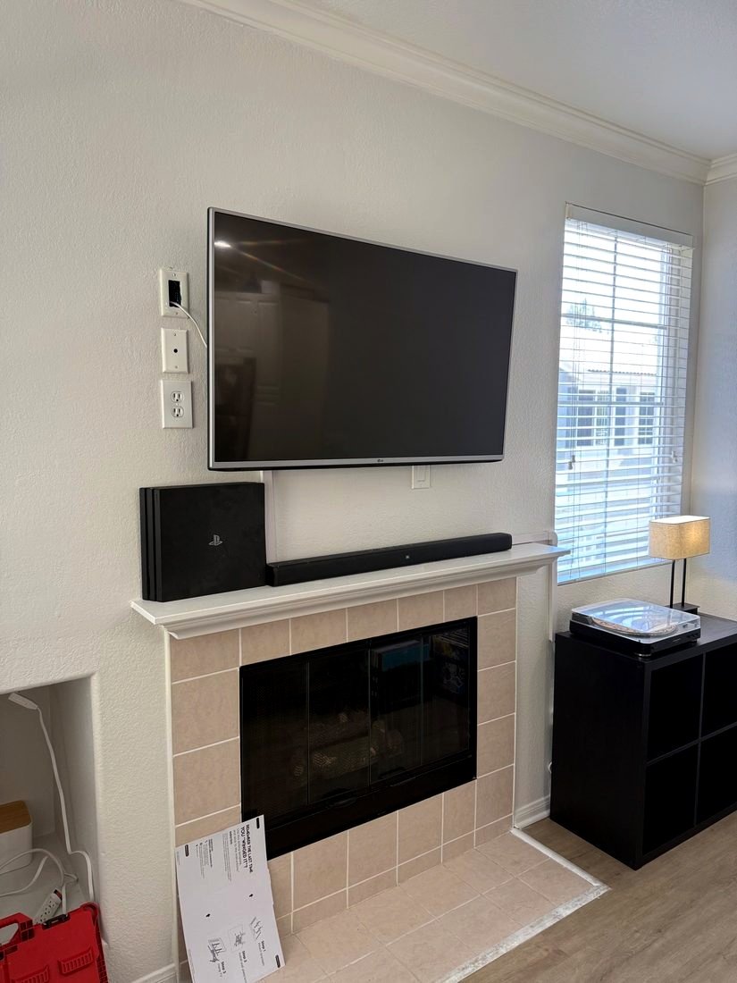 A TV mounted above a tiled fireplace with a soundbar and game console on the mantel